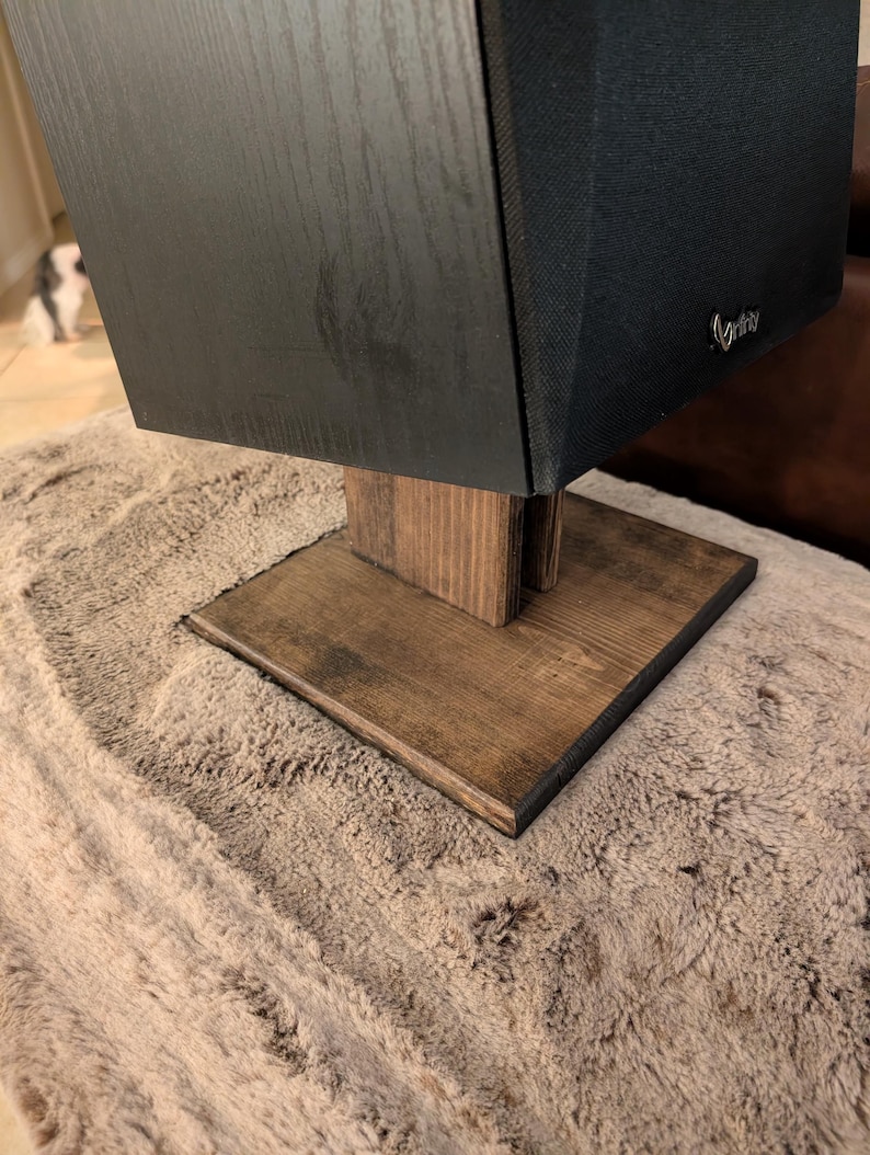 Op de afbeelding: Een zwarte luidspreker met een stoffen grille en een klein zilverkleurig logo staat op een donkerbruine houten standaard. De standaard heeft een vierkante voet en een verticaal steunstuk voor een stabiel platform voor de audioapparatuur.