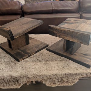 Op de afbeelding: Twee bijpassende luidsprekerstandaards gemaakt van donker gebeitst hout. Elke standaard heeft een platte vierkante voet, een centrale verticale steunpilaar en een schuin geplaatst bovenplatform met een kleine opstaande rand aan de voorzijde.