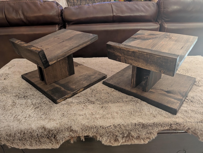 Op de afbeelding: Twee bijpassende luidsprekerstandaards gemaakt van donker gebeitst hout. Elke standaard heeft een platte vierkante voet, een centrale verticale steunpilaar en een schuin geplaatst bovenplatform met een kleine opstaande rand aan de voorzijde.