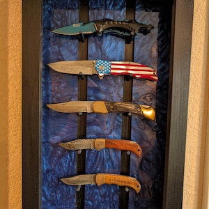 May include: A display case with five knives. The top knife has a teal blade and black handle. The second knife has an American flag design. The third knife has a brown handle. The fourth knife has a wooden handle. The bottom knife has the text "I LOVE YOU".