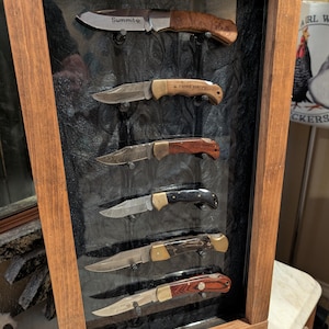 May include: A wooden display case with six pocket knives. The knives have various handle materials, including wood and black composite. The blades are silver, and some have text that reads "Summit" and "I LOVE YOU". The case has a dark background.