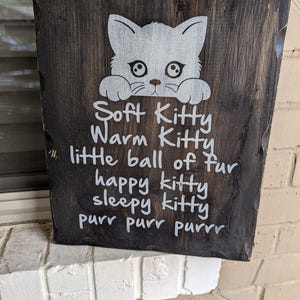 May include: A dark brown wooden sign with white painted text and a white silhouette of a cat peeking over the top. The text reads "Soft Kitty Warm Kitty little ball of fur happy kitty sleepy kitty purr purr purrr".