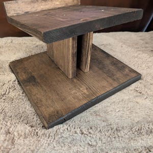 Op de afbeelding: Een donkerbruin gebeitste houten standaard met een vierkante voet, een centrale verticale steunkolom en een vierkant bovenplatform met een kleine achterrand. De houtnerf is over de gehele structuur zichtbaar.