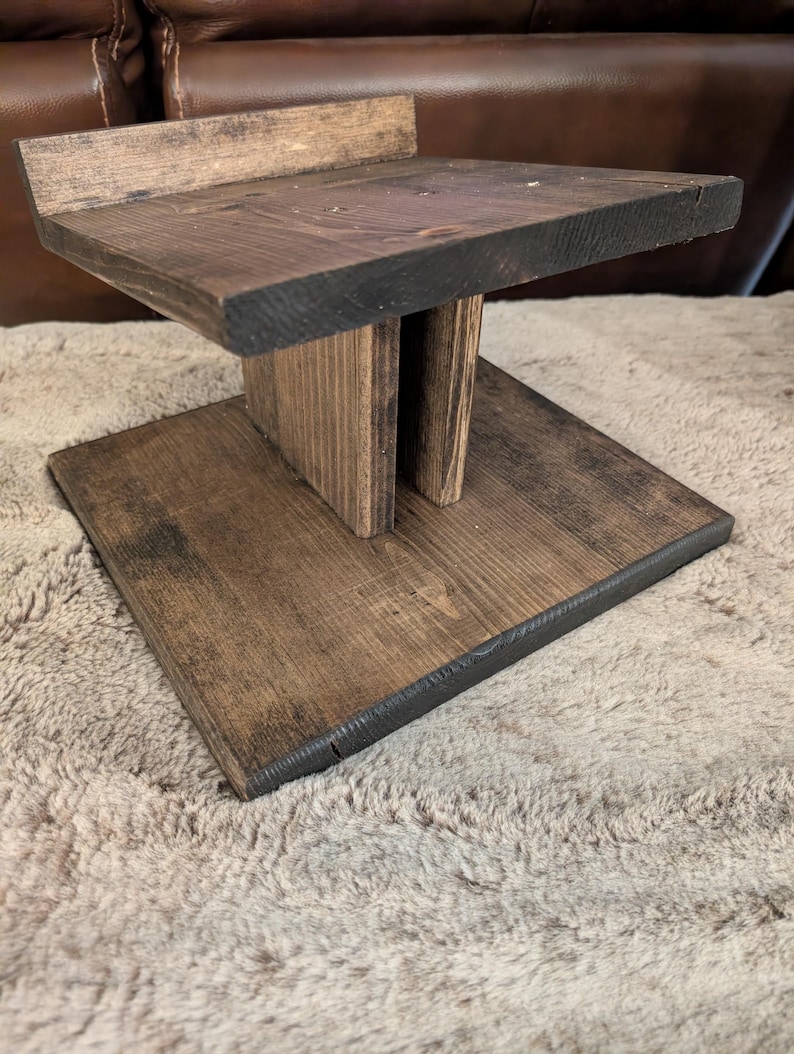 Op de afbeelding: Een donkerbruin gebeitste houten standaard met een vierkante voet, een centrale verticale steunkolom en een vierkant bovenplatform met een kleine achterrand. De houtnerf is over de gehele structuur zichtbaar.