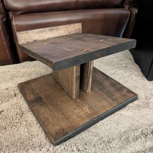 Op de afbeelding: Een donkerbruin gebeitste houten standaard met een vierkante voet, een centrale verticale steunpilaar en een plat bovenplatform met een kleine achterrand. De houtnerf is zichtbaar door de hele structuur en toont een natuurlijke afwerking.