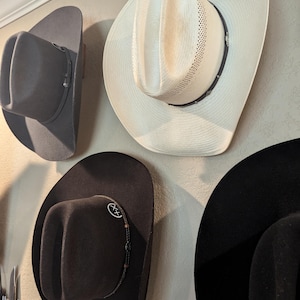 K&ouml;nnte beinhalten: Vier Cowboyh&uuml;te h&auml;ngen an einer Wand. Ein Hut ist wei&szlig;, einer ist grau und zwei sind schwarz. Die schwarzen H&uuml;te haben ein dekoratives Band um den Rand.