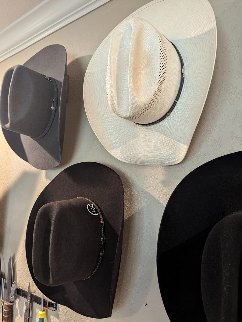 K&ouml;nnte beinhalten: Vier Cowboyh&uuml;te h&auml;ngen an einer Wand. Ein Hut ist wei&szlig;, einer ist grau und zwei sind schwarz. Die schwarzen H&uuml;te haben ein dekoratives Band um den Rand.