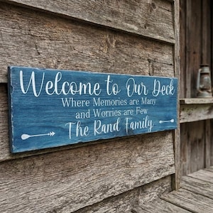 May include: A rectangular blue wooden sign with white text that reads Welcome to Our Deck, Where Memories are Many and Worries are Few, The Rand Family. The sign features two white arrow graphics pointing outward on the bottom line.