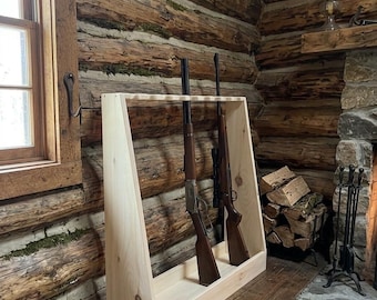 Puede incluir: Un armero de madera de color claro con un marco trapezoidal y un riel superior ranurado diseñado para sostener varias armas largas en posición vertical. El soporte cuenta con una base plana y dos paneles laterales, mostrando dos rifles.