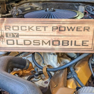 May include: A wooden sign with the text "Rocket Power by Oldsmobile" in black lettering on a brown background.
