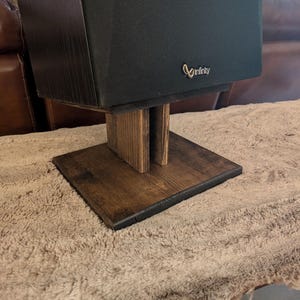 May include: A black Infinity brand speaker mounted on a dark brown stained wooden stand. The stand features a flat rectangular base and two vertical support pillars that elevate the speaker. The speaker has a black fabric grille cover.