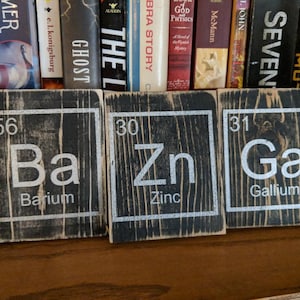 May include: Three wooden signs with black backgrounds and white lettering. The signs are labeled with the chemical symbols and names for Barium (Ba), Zinc (Zn), and Gallium (Ga).