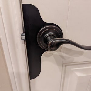 May include: A black door stop with a curved shape is installed on a white door. The door has a bronze door handle with a lever.