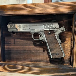May include: A silver Taurus handgun with wooden grips is displayed inside a dark stained wooden shadow box. The shadow box has a rectangular shape with a burnt wood grain finish. The handgun is centered in the frame.