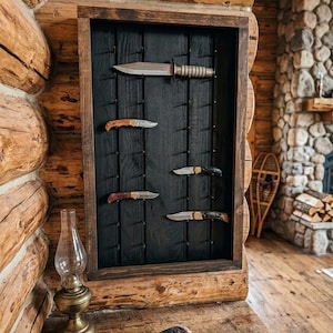May include: A rectangular wooden display case with a dark brown frame and a black vertical-slatted back panel featuring multiple metal pegs. Five various knives with metallic blades and wooden or dark handles are mounted on the pegs in the display.