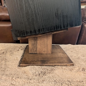 Op de afbeelding: Een zwarte houten luidsprekerkast rust op een donkerbruin gebeitste houten standaard. De standaard heeft een platte vierkante voet, een verticale steunkolom en een schuin geplaatst bovenplatform dat de luidspreker naar boven kantelt.