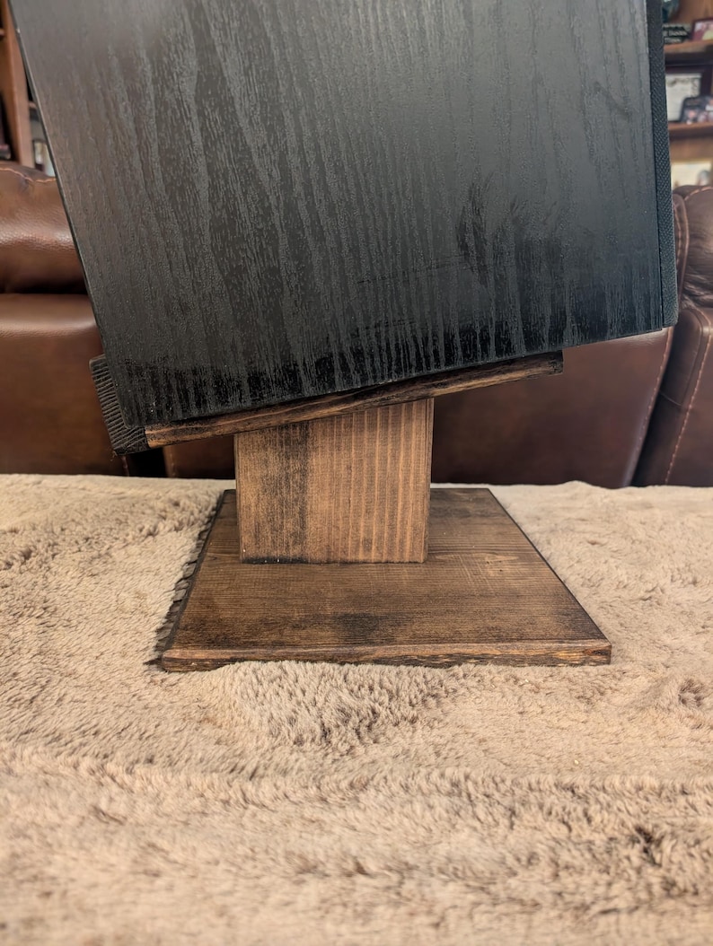 Op de afbeelding: Een zwarte houten luidsprekerkast rust op een donkerbruin gebeitste houten standaard. De standaard heeft een platte vierkante voet, een verticale steunkolom en een schuin geplaatst bovenplatform dat de luidspreker naar boven kantelt.