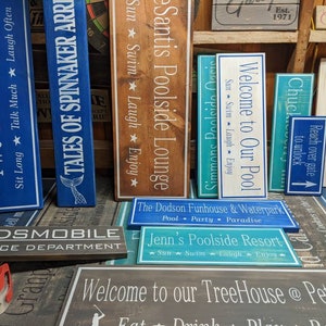 May include: A collection of wooden signs with various text and designs. The signs are painted in blue, white, brown, and green. The text on the signs includes "Two Bunch Grill", "Tales of Spinnaker Arri", "Santis Poolside Lounge", "Welcome to Our Pool", "Welcome to Our Patio", "The Dodson Funhouse & Waterpark", "Jenn's Poolside Resort", and "Welcome to our TreeHouse @ Pete Dye".