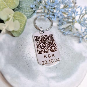May include: Silver keychain with a black and white QR code, the text "K & K" and the date "22.10.24" engraved on it.