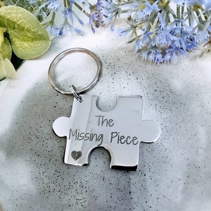 Może przedstawiać: Srebrny brelok do kluczy w kształcie puzzla z wygrawerowanym napisem "The Missing Piece". Po lewej stronie puzzla wygrawerowane jest małe serce.