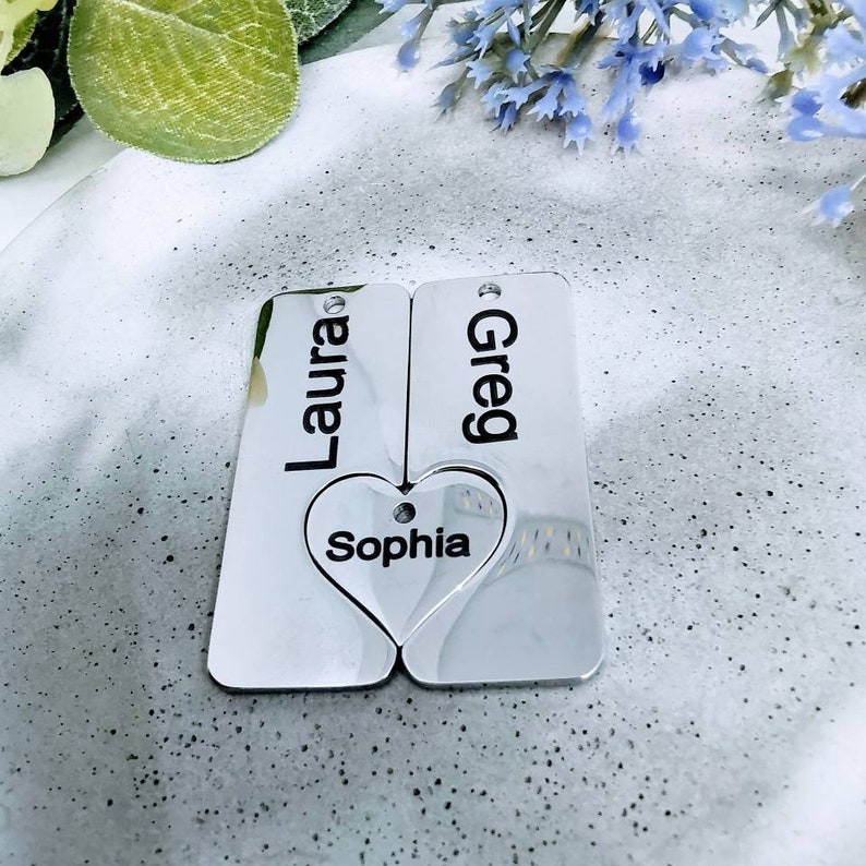 May include: Two silver rectangular tags with the names "Laura" and "Greg" engraved on them. A heart-shaped tag with the name "Sophia" engraved on it is attached to the tags.