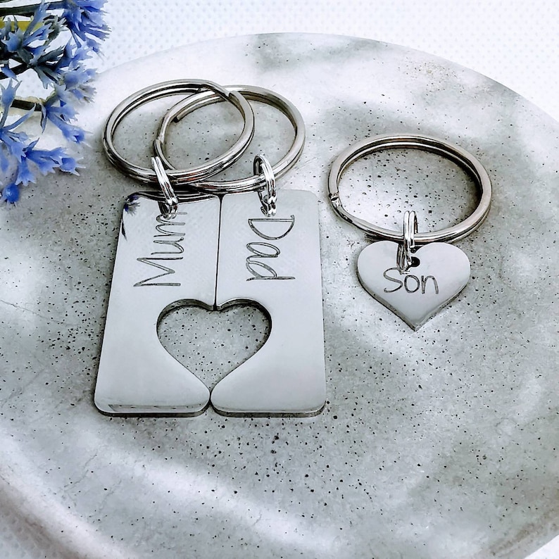 May include: A set of three silver keychains with engraved words "Mum", "Dad", and "Son". The "Mum" and "Dad" keychains are connected by a heart shape cutout.