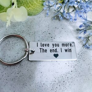 Puede incluir: Llavero de plata con una etiqueta rectangular que dice "Te amo más. El final. Yo gano" con un pequeño símbolo de corazón al final.