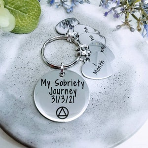 Puede incluir: Llavero de plata con un dije redondo que dice "My Sobriety Journey 3/3/21" con un símbolo de triángulo. El llavero también tiene varios dijes en forma de corazón con las palabras "1 Month", "3 Months" y "6 Months" en ellos.