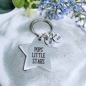 May include: A silver keyring featuring a star-shaped charm inscribed with "POPS' LITTLE STARS." Two small, round charms are attached, one with "CHLOE" and the other with "ALEX." The keyring rests on a grey surface with blue flowers.