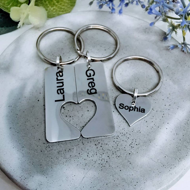 May include: Two silver keychains with the names "Laura" and "Greg" engraved on them. The keychains are connected by a heart shape. A third silver keychain with a heart shape and the name "Sophia" engraved on it.