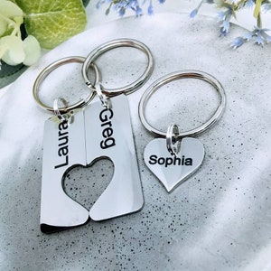 Puede incluir: Llaveros de plata con dijes de corazón entrelazados y etiquetas con nombres personalizados. Las etiquetas dicen "Laura", "Greg" y "Sophia".