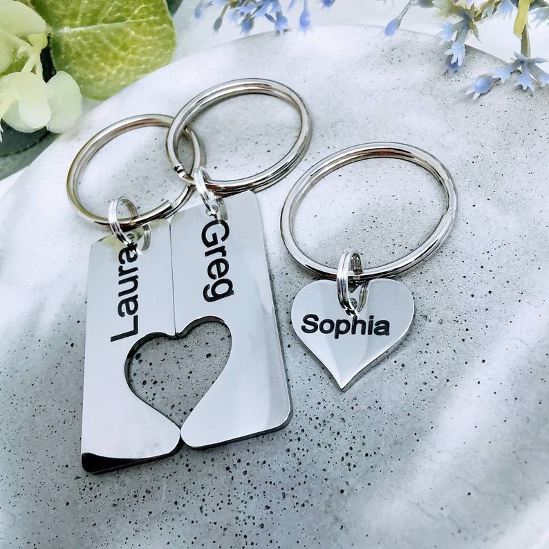 May include: Silver keychains with interlocking heart charms and personalized name tags. The tags read "Laura", "Greg", and "Sophia".
