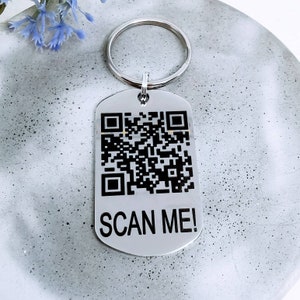 Op de afbeelding: Zilveren sleutelhanger met een zwart-witte QR-code en de tekst "SCAN ME!" erop.
