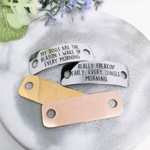 Puede incluir: Tres chapas de perro de metal con diferentes lemas. La chapa superior es de plata y dice "Mis perros son la razón por la que me despierto cada mañana". La chapa del medio es de plata y dice "Realmente temprano. Cada. Mañana.". La chapa inferior es de oro.