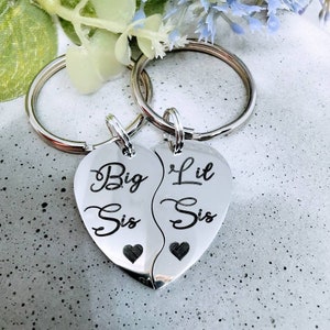 Op de afbeelding: Twee zilveren hartvormige sleutelhangers met de woorden "Big Sis" en "Lil Sis" erop gegraveerd. De sleutelhangers zijn aan elkaar verbonden en hebben kleine hartjes aan de onderkant.