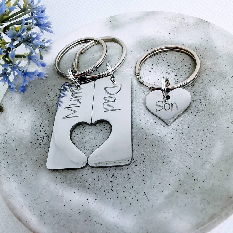 May include: Two silver keychains with interlocking heart cutouts. One keychain says "Dad" and the other says "Mum". A third silver keychain with a heart charm says "Son".