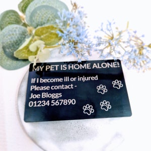 Pode incluir: Etiqueta de metal preto com texto branco que diz "MY PET IS HOME ALONE! If I become ill or injured Please contact - Joe Bloggs 01234 567890". Há três pegadas de patas na etiqueta.