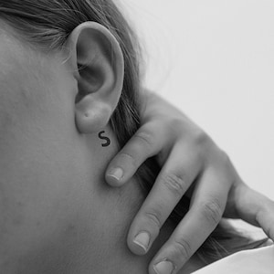 Peut inclure: Gros plan d'une personne portant une chemise blanche à col. La personne a un petit tatouage de la lettre "S" derrière l'oreille.