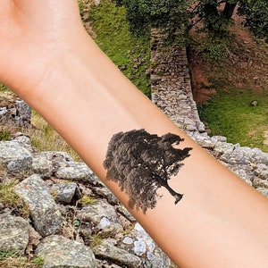 Puede incluir: Un brazo con un tatuaje en blanco y negro de un árbol en la muñeca. El tatuaje está en primer plano de la imagen, con una colina verde cubierta de hierba y un muro de piedra en el fondo.