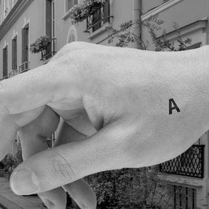 May include: A black and white photograph of a narrow street in a city. The street is lined with buildings on both sides. A hand is pointing down the street, with the letter "A" tattooed on the back of the hand.