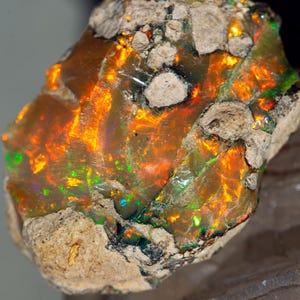 Könnte beinhalten: Ein roher, natürlicher Opal mit einem leuchtenden orangefarbenen, grünen und blauen irisierenden Schimmer. Der Opal ist in eine Matrix aus braunem und grauem Gestein eingebettet.