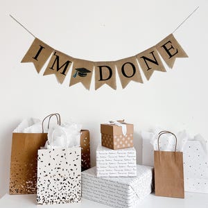 May include: A burlap banner with the text "I'M DONE" in black lettering. The banner has a graduation cap icon in the middle. The banner is hanging on a white wall above a table with brown and white gift bags and boxes.