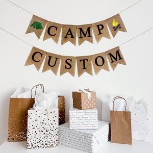 A custom burlap banner made from 4.5 by 6.5 inch fishtail shaped flags. Each flag has a pocket across the top for stringing. The top line of this banner says CAMP in black capital letters. There is a hand stenciled image of a green tent on one end and green pine trees on the other end. The second line can be customized with a name in black capital letters. It is strung on two jute strings that are 110 inches in length.