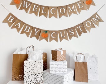May include: Welcome Baby Custom burlap banner with orange pumpkins and white text. The banner is hanging above a table with brown and white gift bags.