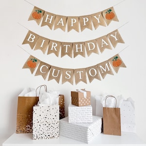 May include: A burlap banner with the words "Happy Birthday Custom" in white letters. The banner is decorated with small orange pumpkins. The banner is hanging on a white wall above brown gift bags.