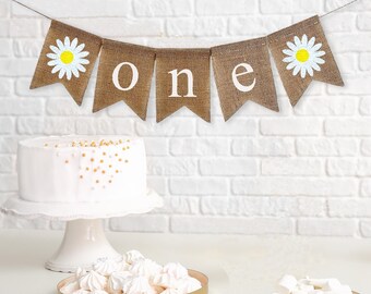 Op de afbeelding: Een jute vlag met witte madeliefjes en het woord "one" in witte letters. De vlag is aan een witte muur bevestigd. Er staat een witte taart en een bord met witte meringue-koekjes op een tafel voor de vlag.