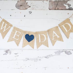 Pu&ograve; includere: Uno striscione di juta con le parole "We Love Dad" in lettere bianche. Un cuore blu &egrave; tra le parole "Love" e "Dad".