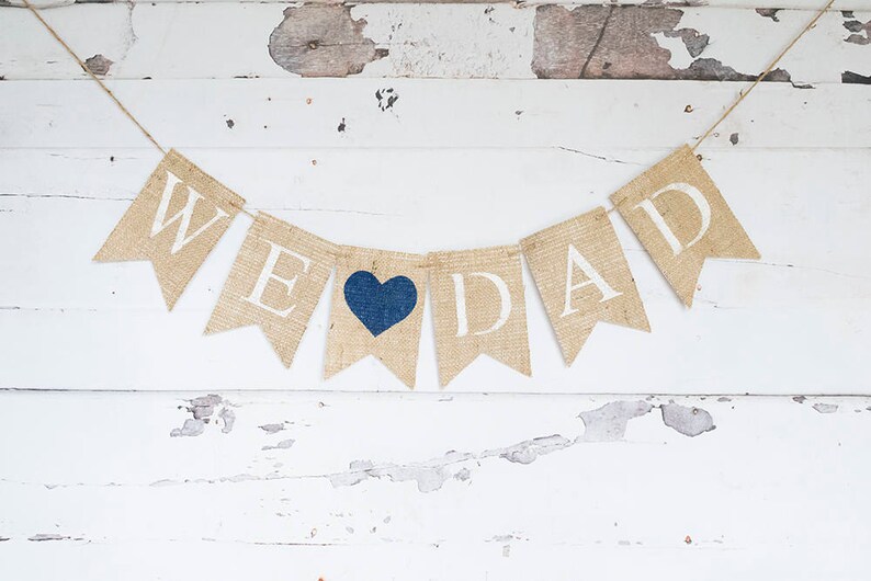 Pu&ograve; includere: Uno striscione di juta con le parole "We Love Dad" in lettere bianche. Un cuore blu &egrave; tra le parole "Love" e "Dad".