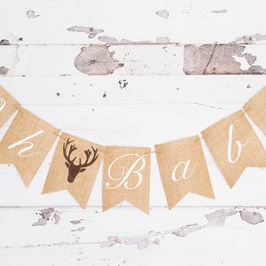 May include: A burlap banner with the words "Oh baby" in white script. The banner has a brown deer silhouette on one of the flags.