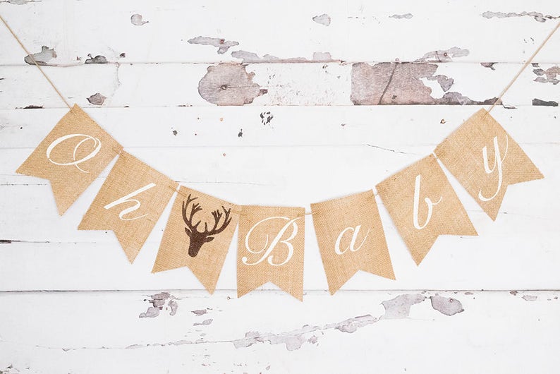 May include: A burlap banner with the words "Oh baby" in white script. The banner has a brown deer silhouette on one of the flags.